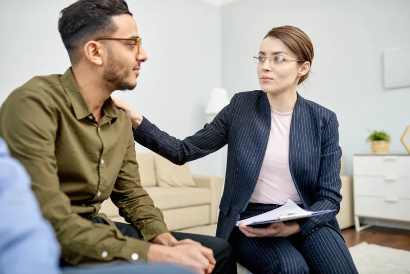 Psychiatric mental health provider comforting a client by placing a hand on his shoulder in a cozy office setting.